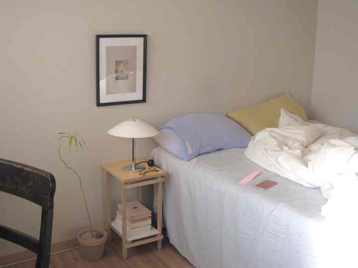 ライトグレーの壁と木目調のナイトスタンドがあるシンプルでナチュラルな一人暮らしの寝室、白い寝具とパステルカラーの枕が置かれたベッドを特徴とする