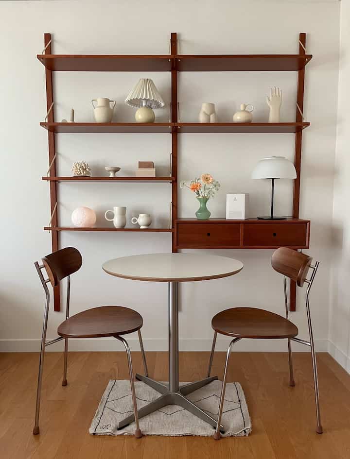 Wood tone and white compact dining area featuring walnut shelves, round table and two chairs in a simple natural interior