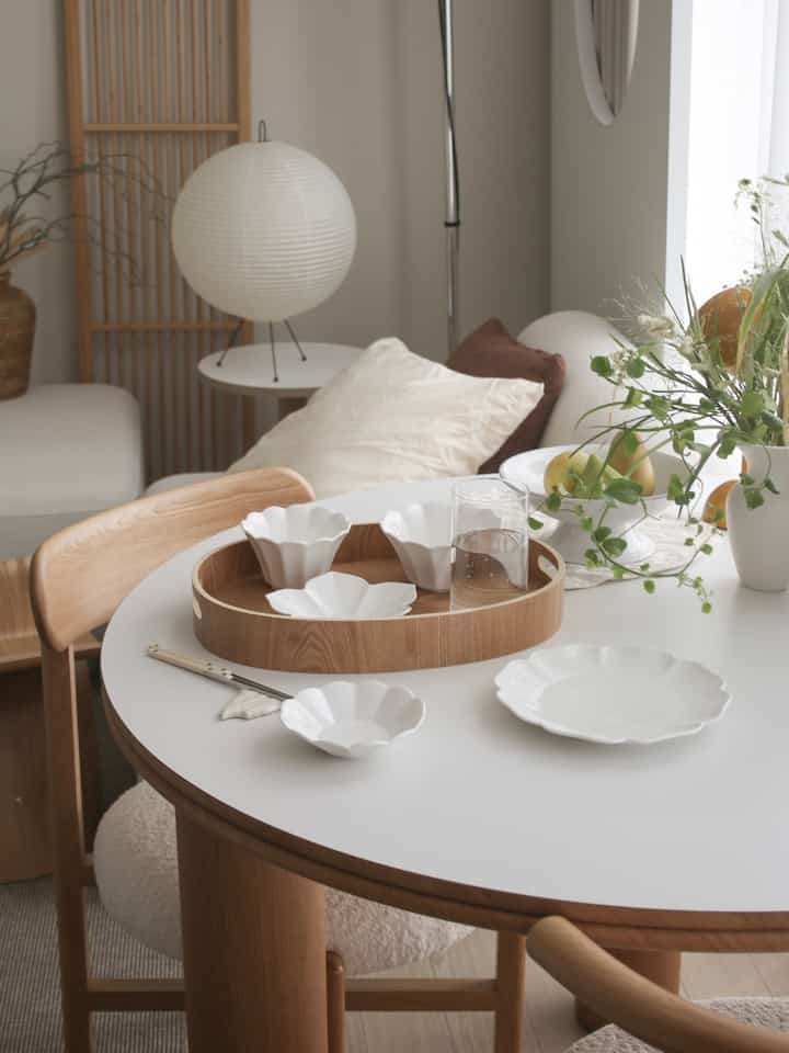Natural-toned two-person dining room featuring a round white dining table, wooden tray, ceramic dishware, and greenery creating a cozy atmosphere