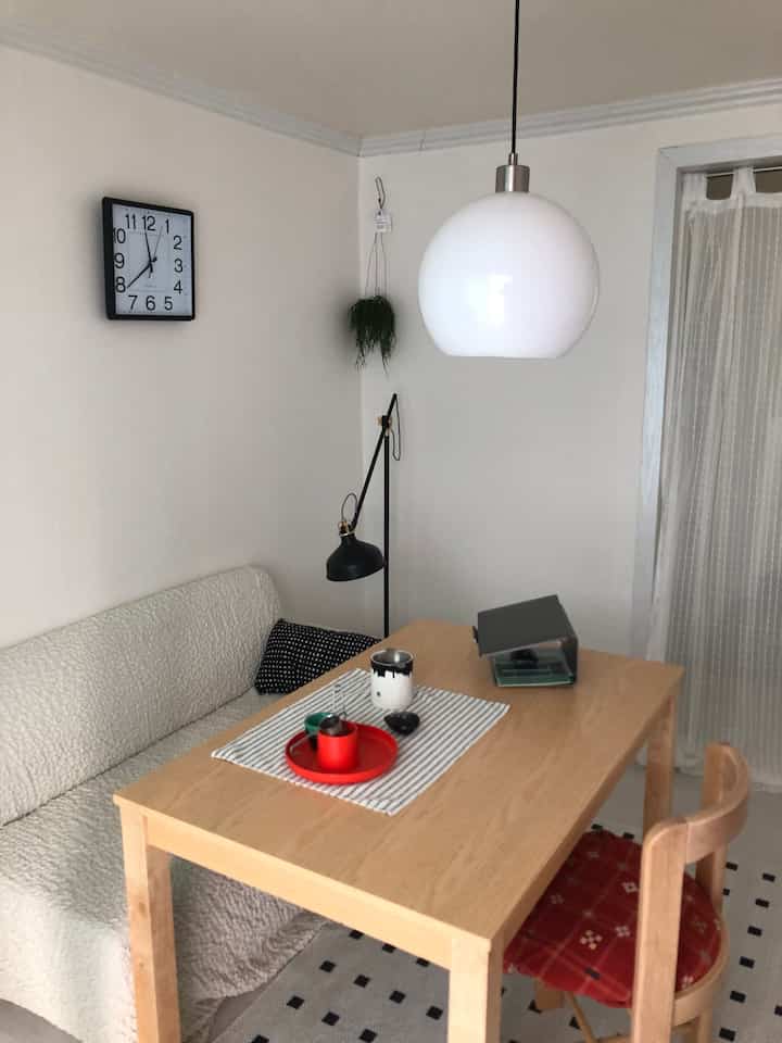 ベージュの壁と木目調のダイニングテーブルがあるシンプルなワンルームリビング、白いソファと赤いクッションの椅子が配置された雰囲気のあるインテリア