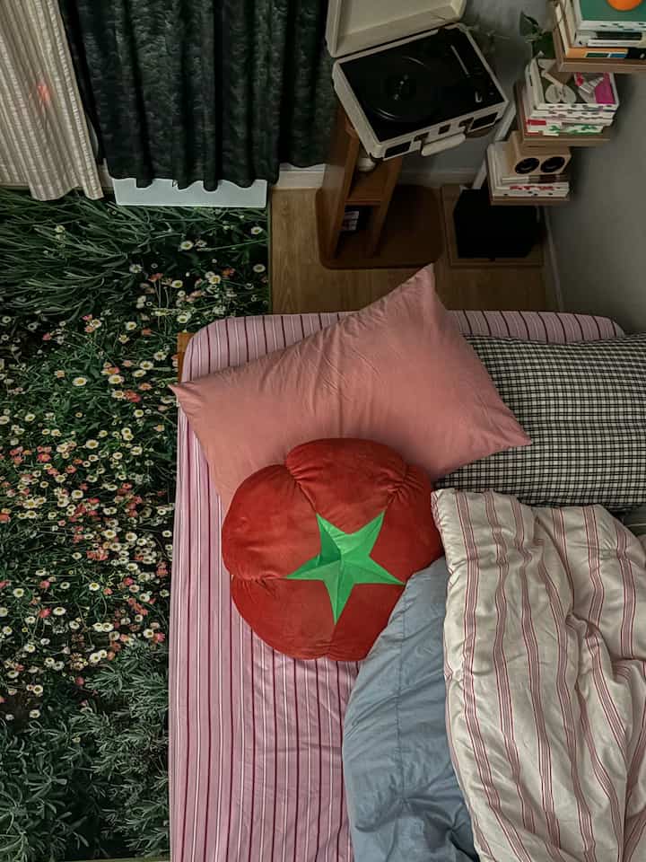 緑のフロアマットと木目調のサイドテーブルがあり、ピンククッションと赤いトマトクッションが置かれた寝室のインテリア