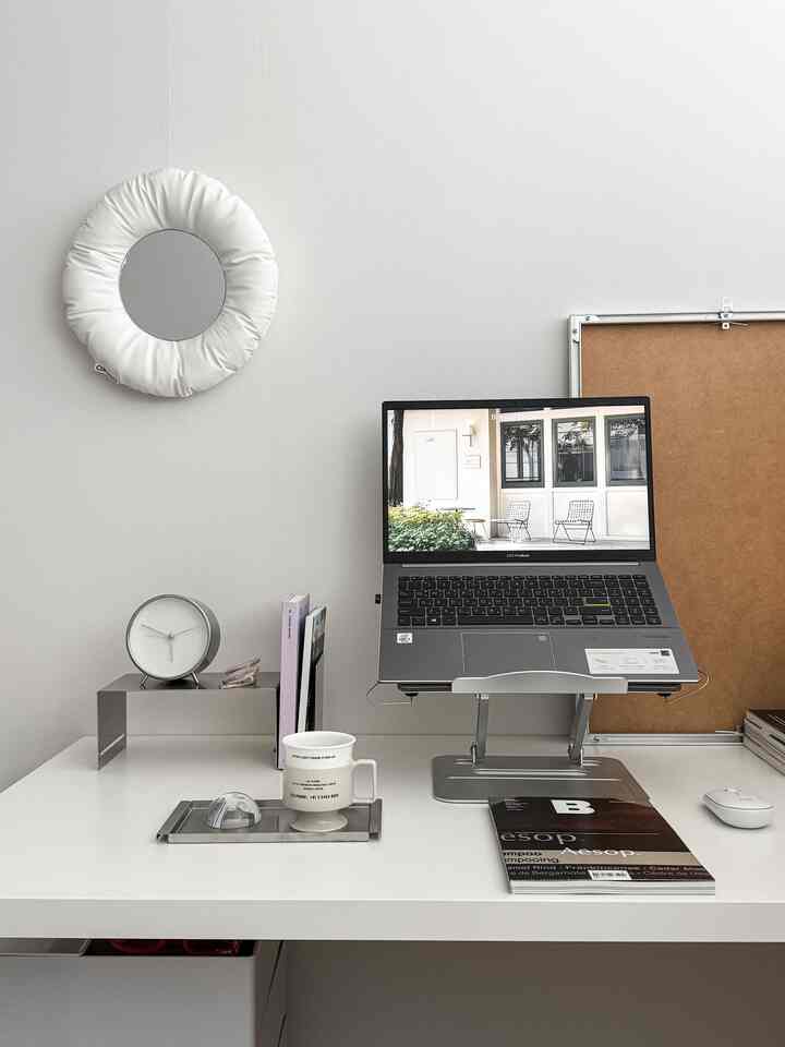 Modern white-toned study featuring a white desk with a laptop stand, clock, and organizer neatly arranged
