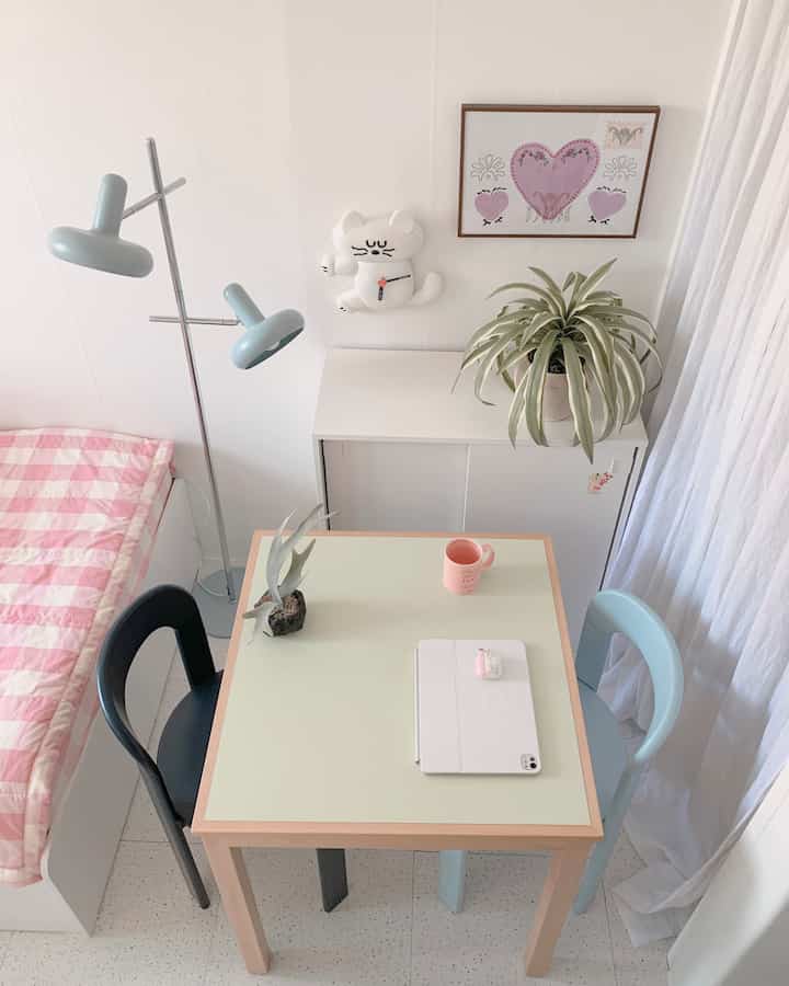 Bright wood-toned studio apartment featuring a light green wood table, pink check bedding, linen curtains, plants, and a cat wall clock creating a cozy cafe-style space
