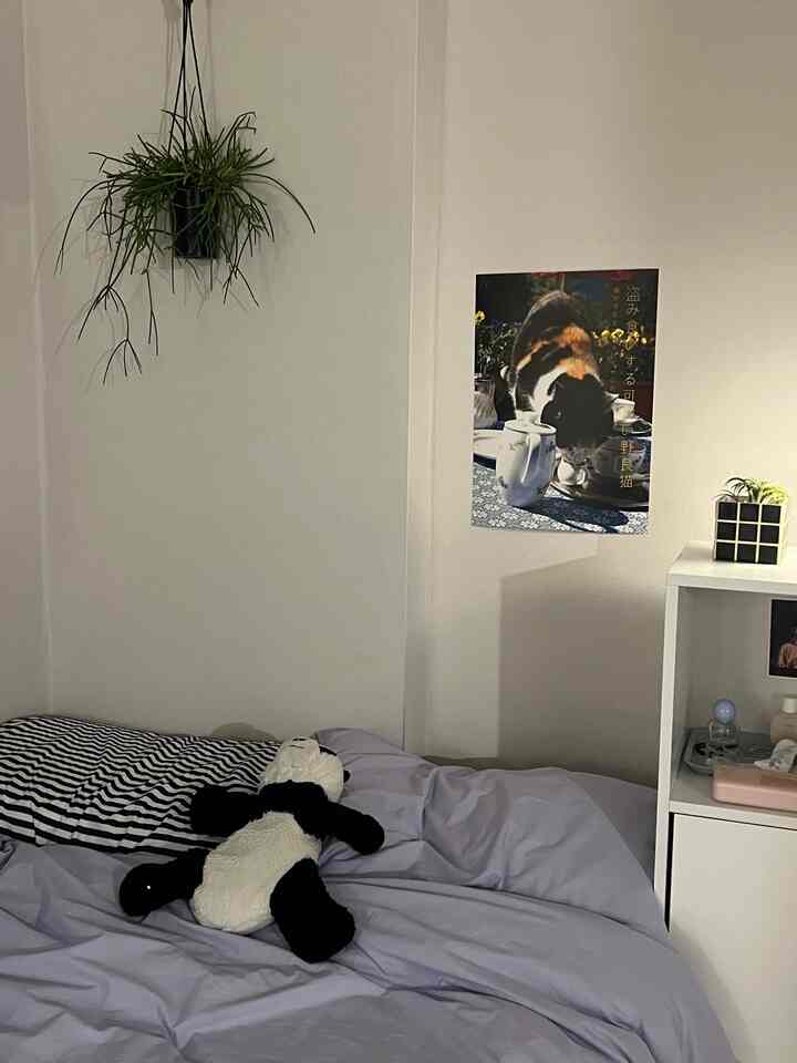 白い壁とラベンダーの掛け布団がある寝室、枕とパンダのぬいぐるみ、壁に猫のポスターと植物が配置された雰囲気のあるインテリア