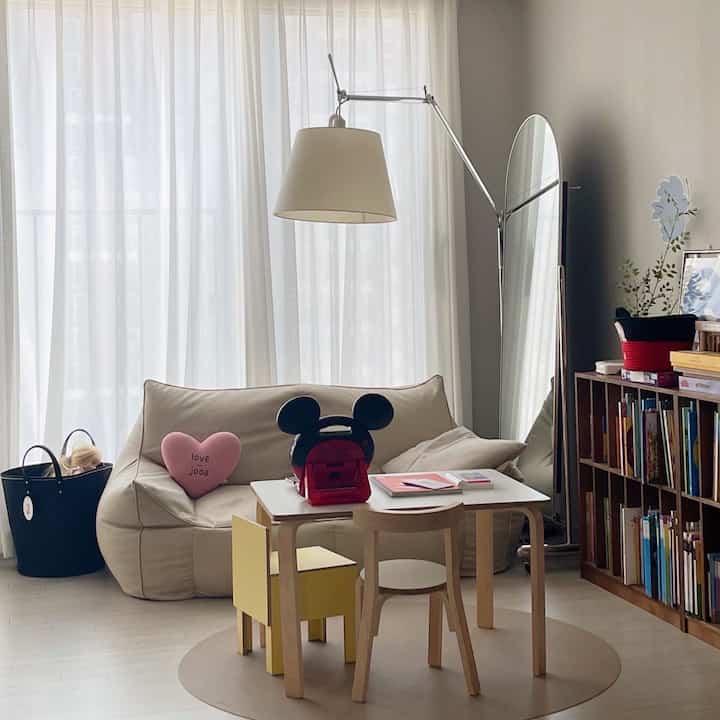 白いカーテンの背景にナチュラルな色調のリビング、小さな子供用机とビーズクッションが配置された赤ちゃんと暮らす快適な空間