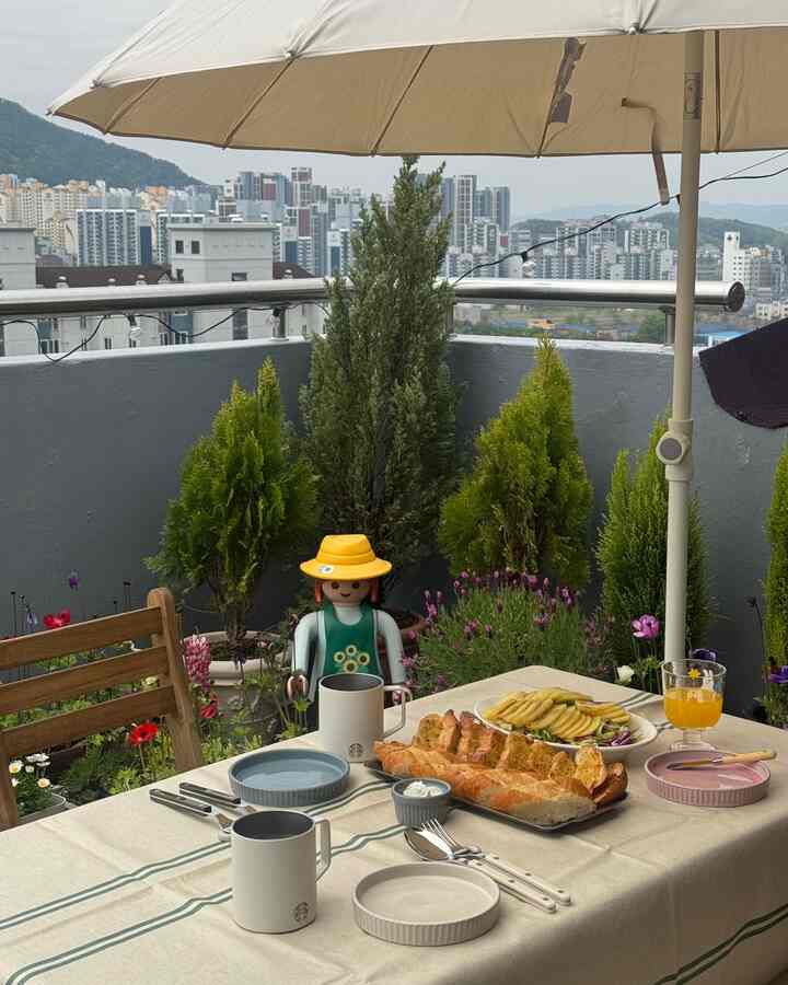 ベージュのパラソルがあるバルコニーにダイニングテーブルと椅子、多様な食器とパン、ドリンクが配置された雰囲気のあるホームカフェ空間