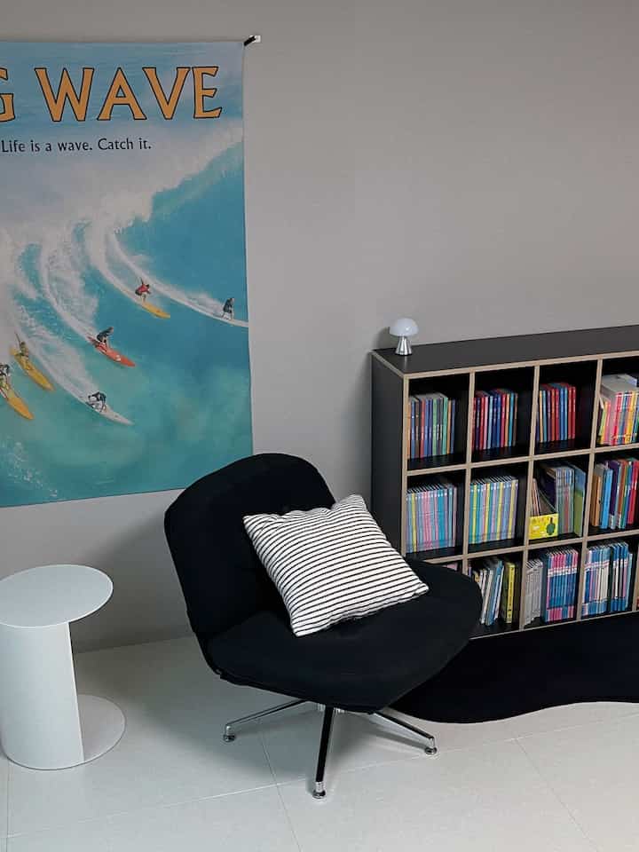 Modern living room with white tiled floor and walls, featuring a black armchair with striped cushion and a black bookshelf filled with colorful children's books