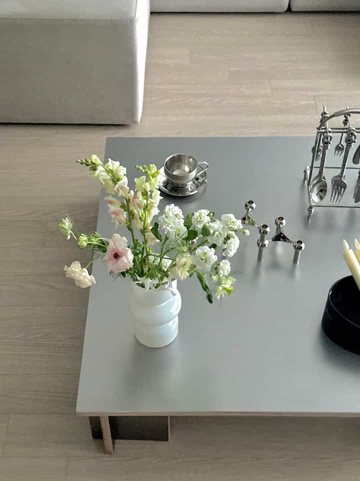 明るい木目の床とシルバーのコーヒーテーブルに白い花瓶と様々な花が置かれたミニマルなリビング空間