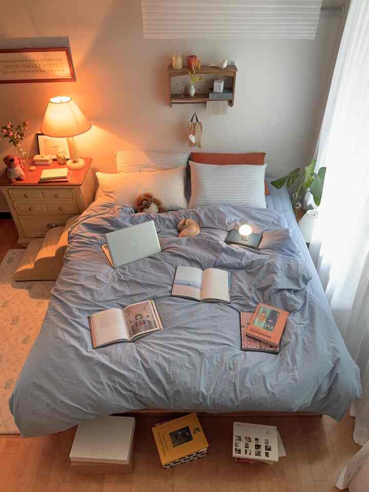 ベージュ色の壁とライトブルーの寝具がある一人暮らしの寝室、本や暖かい照明が配された自然で落ち着いた雰囲気のインテリア