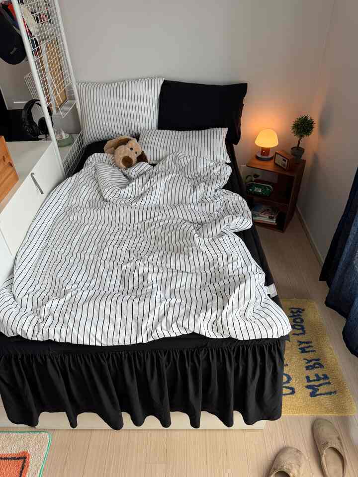 Studio bedroom with black and white striped bedding, elevated bed with bed skirt, nightstand lamp creating cozy atmosphere