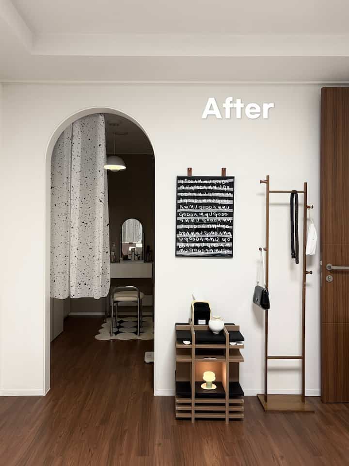 白い壁と木目調の床のワンルームウォークインクローゼット、アーチ型の入口に化粧台とカーテン、壁にポスターと木製コートラックが配置された雰囲気のあるインテリア