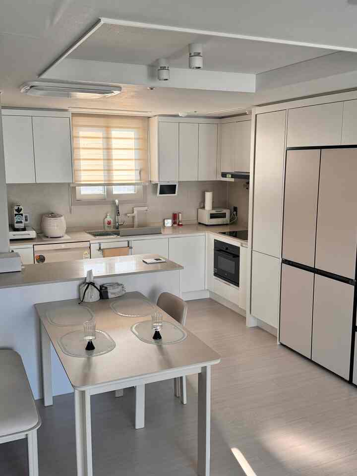 明るいホワイトとベージュ調のキッチン空間、ダイニングテーブルとブラインドが配置され、モダンで自然な雰囲気のインテリア