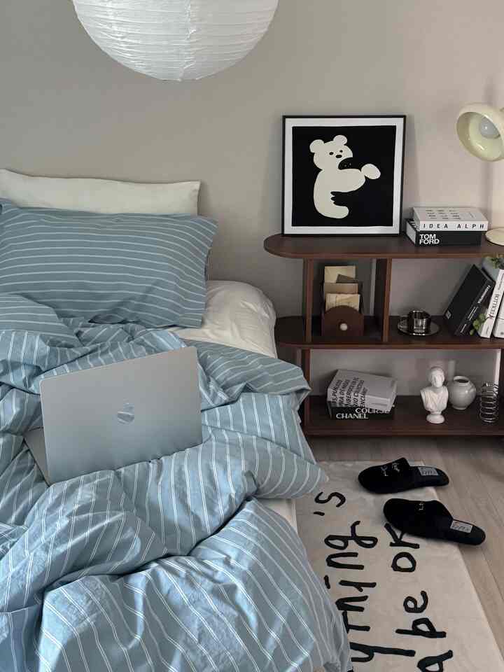 ホワイトの壁とライトグレーの床のナチュラルトーンの寝室、青のストライプ掛け布団カバーと木製の棚が調和した居心地の良い空間