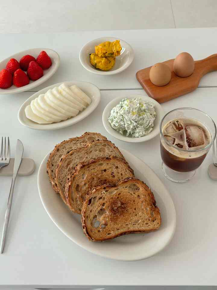 White-toned dining table featuring toasted bread, eggs, strawberries, and iced coffee in a home cafe breakfast setup
