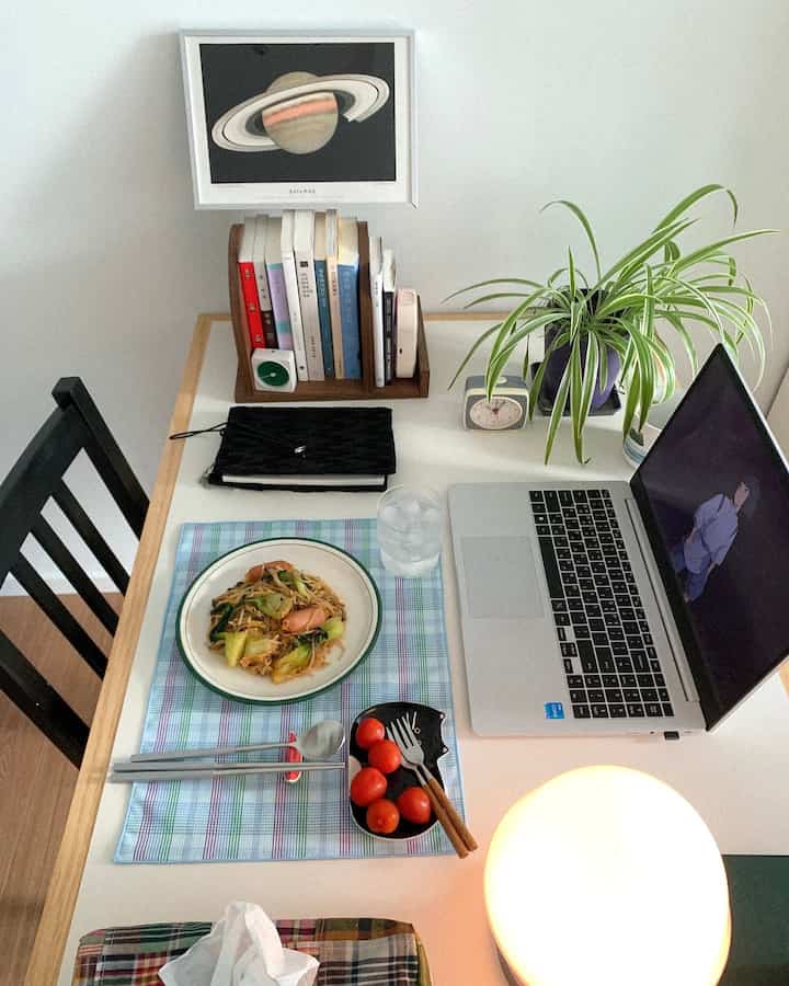 白いテーブルの上にノートパソコン、食器、本棚、観葉植物が配置されたシンプルなホームオフィスの雰囲気のあるインテリア