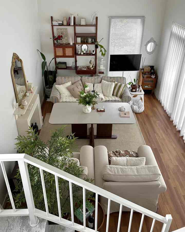 ベージュと木目調のリビング、二つのソファと中央のテーブル、植物と壁棚が配置された自然光あふれるナチュラルモダンなインテリア