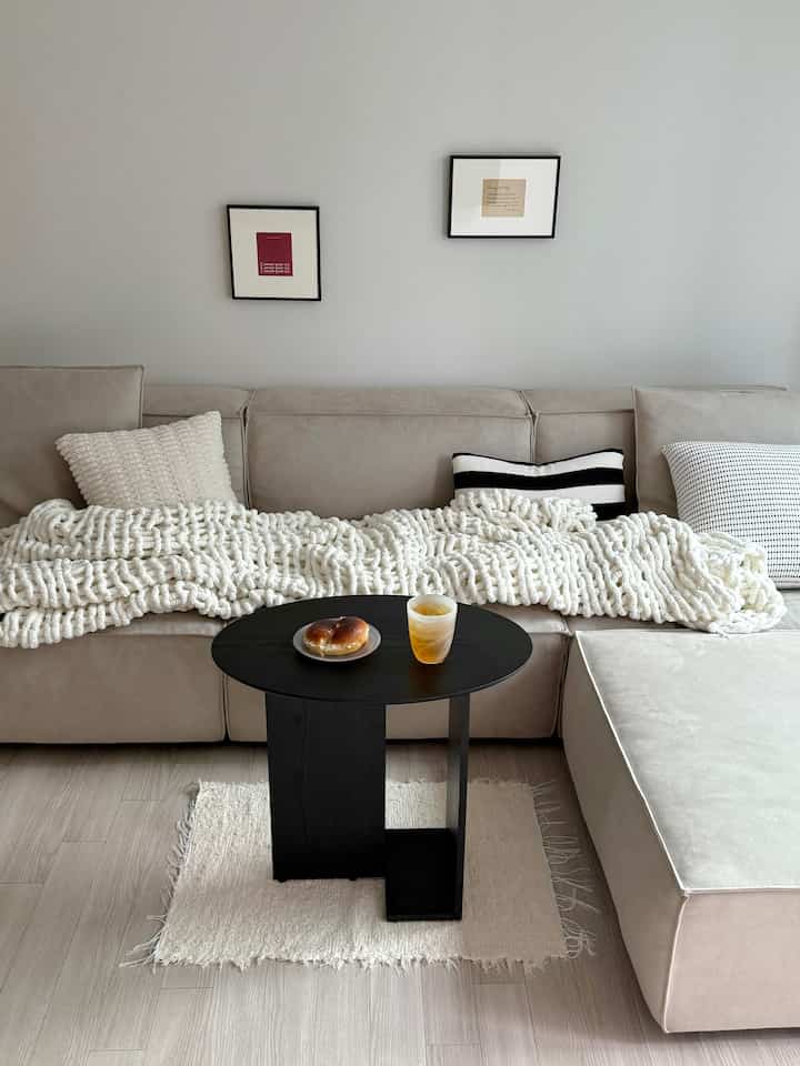 Beige and white toned living room with beige sectional sofa draped in chunky white knit blanket and a black round coffee table, creating a modern atmosphere