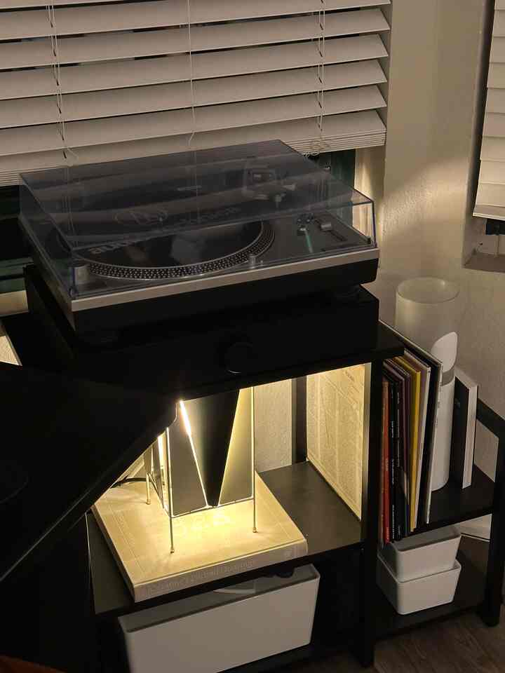 Black-toned corner shelving featuring a table lamp and turntable, creating a cozy minimal music listening area with warm ambient lighting