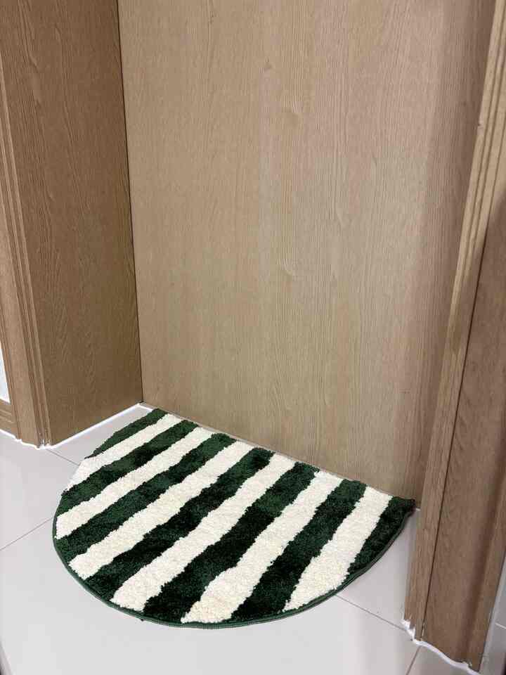 淡い木目調の壁と白いタイル床に配置された、深緑と白のストライプの半円形ドアマットがあるキッチン入口の空間