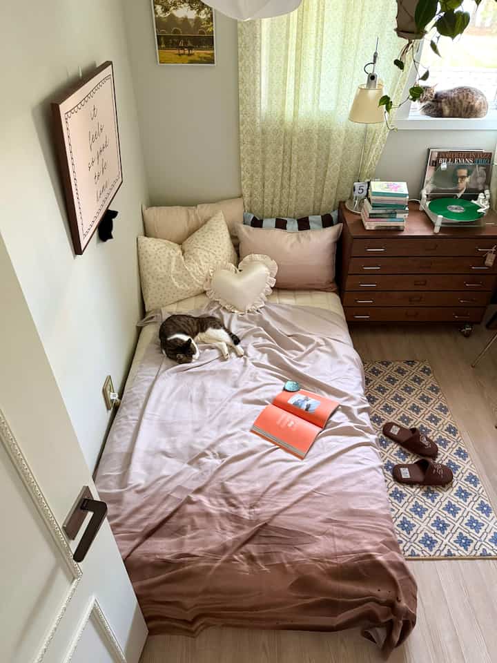 ベージュとブラウンのトーンの寝室、クッションの上でくつろぐ猫がいるベッドと、窓際にある木目調のチェストが温かみのある雰囲気を演出する空間
