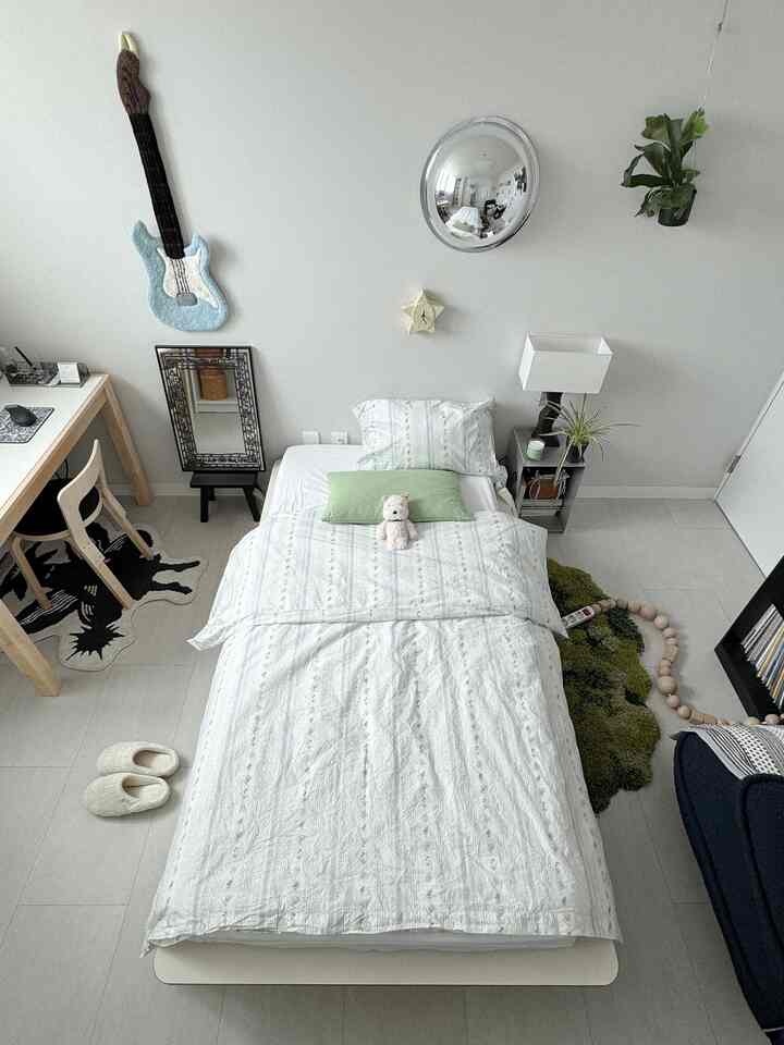 ホワイトとグリーンのトーンのワンルーム寝室、中央にシングルベッドと枕、壁に丸い鏡と植物が配置されたミッドセンチュリーモダンなインテリア