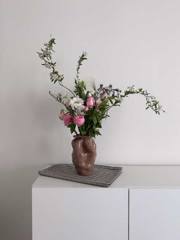 モノトーンの白い寝室、中央に花瓶と花束が置かれた白いキャビネットが配置されたシンプルで自然な雰囲気のインテリア