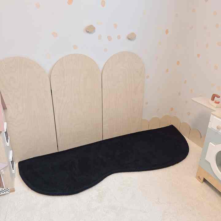 ナチュラルな木目調の子供部屋、アーチ型の木製パネルと黒いラグが配置されたシンプルで落ち着いた空間