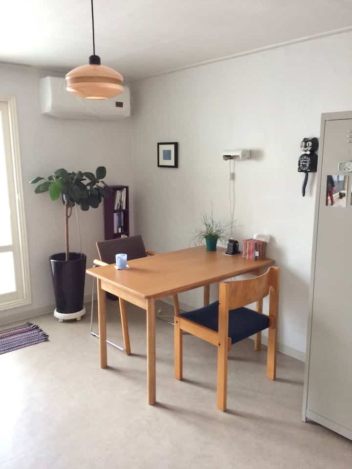 白い壁と木目調のダイニングテーブルが配置されたシンプルで狭いダイニング、植物と壁掛け時計がある雰囲気の良いインテリア