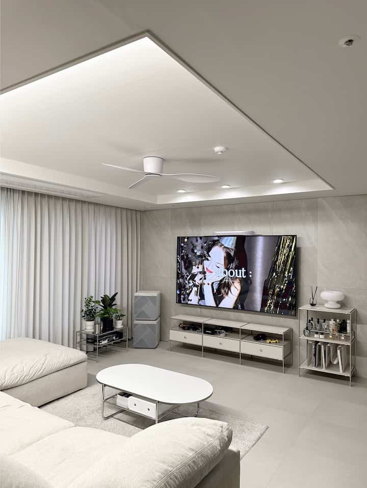 明るいホワイトとグレーのモノトーンリビング、大きなソファと白いコーヒーテーブル、壁掛けテレビとシーリングファンが配置された清潔でモダンな空間