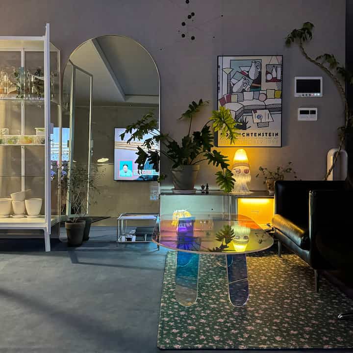 グレーの壁とネイビーフロアのモダンなリビング、中央に透明なコーヒーテーブルとソファ、植物とテーブルランプが配置された雰囲気のあるインテリア