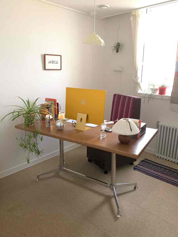 ベージュのタイルカーペットと木製デスクが配置されたナチュラルな書斎、黄色のアイマックと植物が調和するシンプルなホームオフィスのインテリア