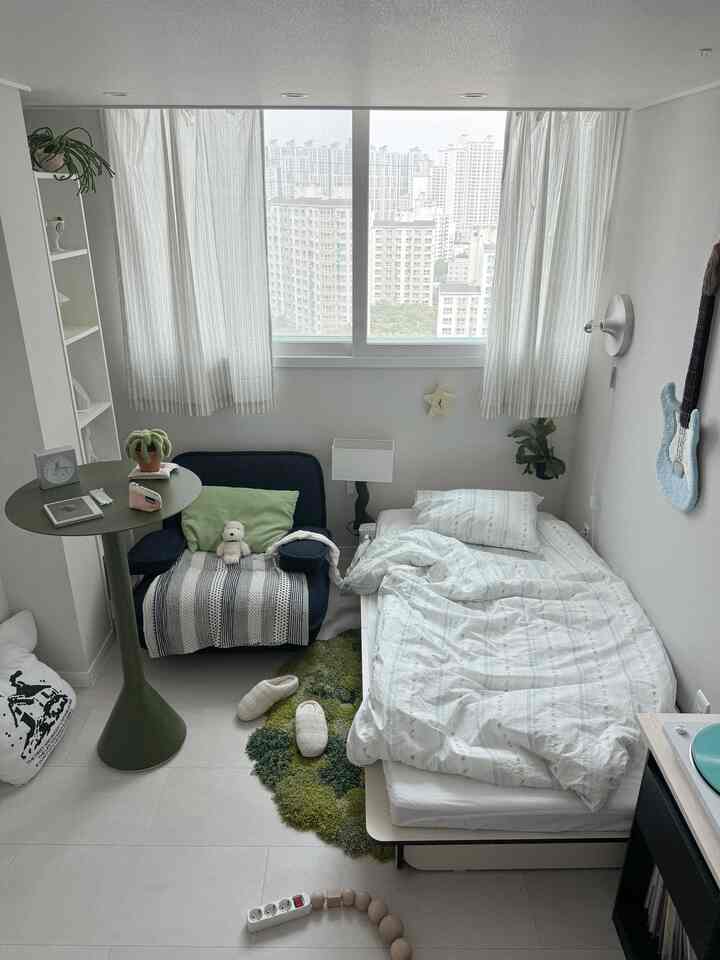 明るいナチュラルトーンのワンルーム寝室、ネイビーのアームチェアとオリーブグリーンのスタンディングテーブルが配置されたシンプルで居心地の良い空間