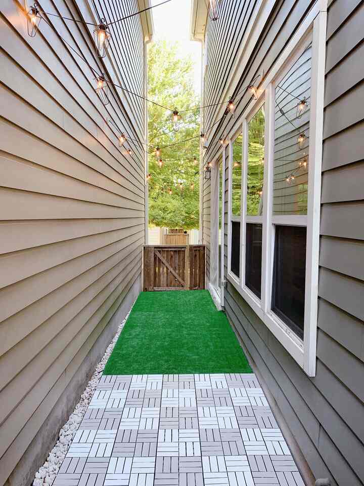 Natural-toned narrow veranda featuring artificial grass rug, decking tiles, and vintage string lights for a cozy outdoor space