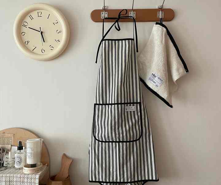 アイボリーの壁にクリーム色の時計とストライプのエプロン、タオルが掛かるナチュラルでシンプルなキッチンスペース