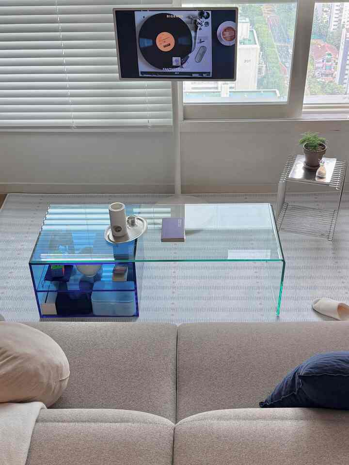 ベージュのソファと透明なガラスのコーヒーテーブルが配置されたモダンなリビング、白いモニタースタンドと青のアクセントカラーのある空間