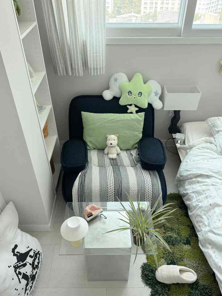 Compact modern living room with navy single sofa featuring green pillow, white nightstand, and visible bed under natural light