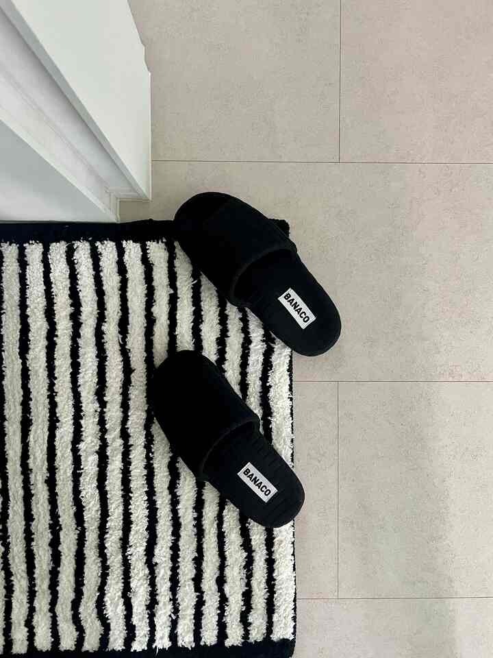 ブラックとホワイトのコントラストが際立つバスマットの上に置かれた黒い室内履きがある印象的な玄関前の空間