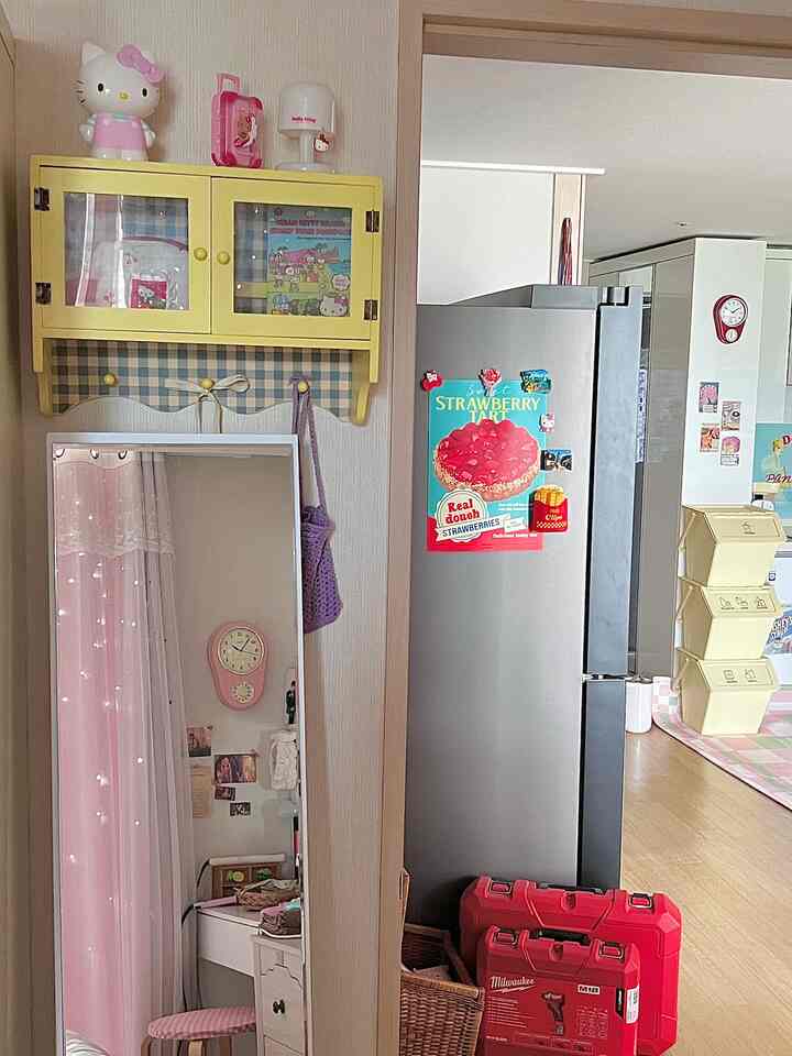 ピンクとヴィンテージ調の寝室で、黄色い壁棚とピンクの時計、カーテン、冷蔵庫が配置された雰囲気のあるインテリア