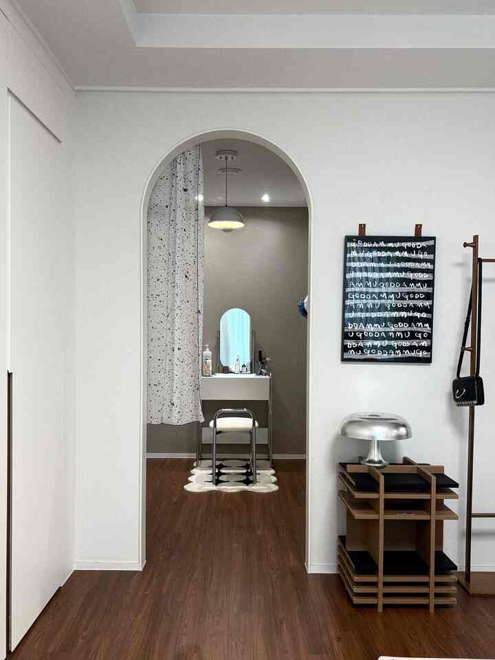 白と木目調のトーンが特徴のアーチゲートのあるパウダールーム、スリムな化粧台と楕円形の鏡、白いスツールが配置されたシンプルでモダンな空間