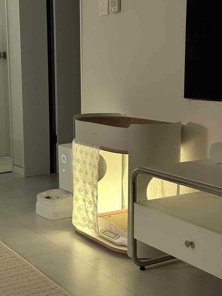 ホワイトとナチュラルカラーのリビング、あたたかな間接照明に照らされた猫用スクラッチャーと自動給餌機が配置された雰囲気のあるインテリア