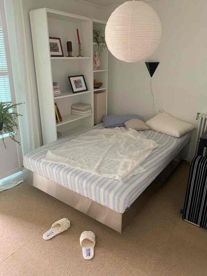 白とナチュラルな色調の寝室、シンプルなベッドフレームと本棚、居心地の良い寝具と壁照明が配置された印象的な空間