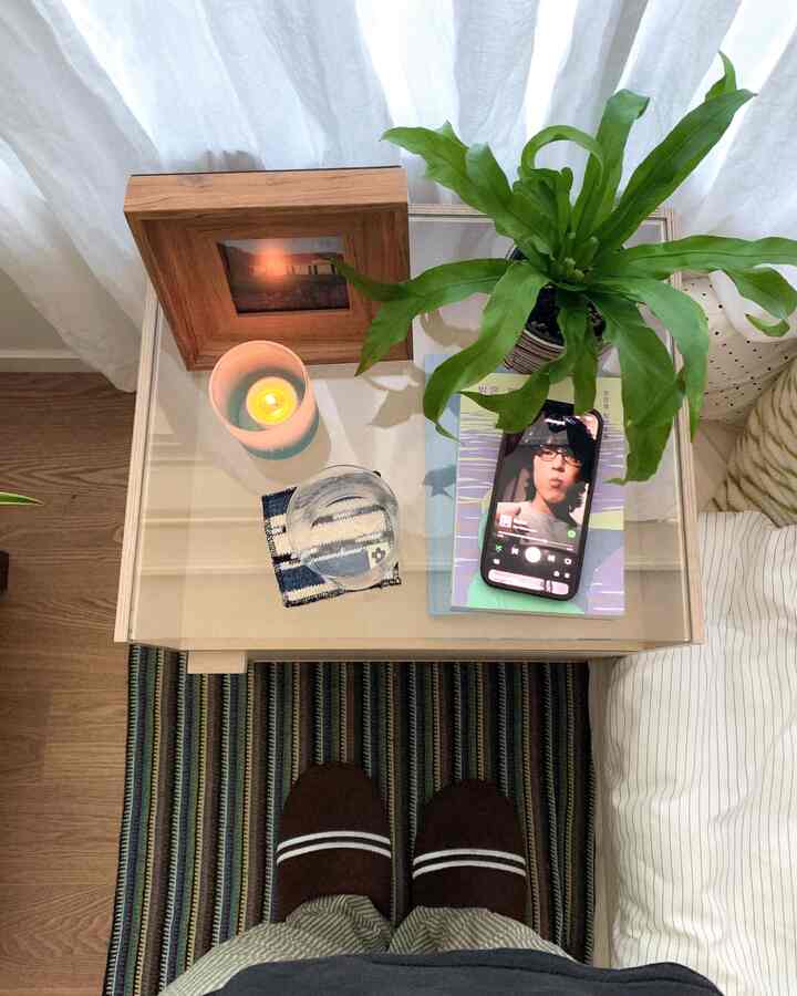 明るいベージュ調の小さな寝室のナイトテーブルに緑の植物とキャンドル、写真立てが置かれた落ち着いた空間