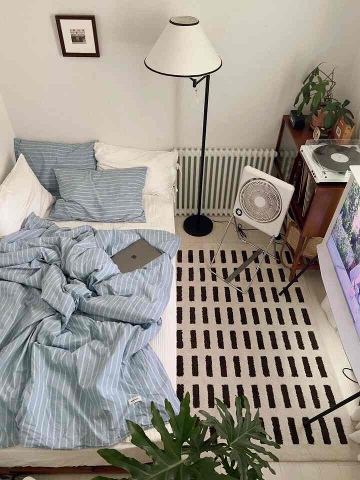 白い壁と青いストライプの寝具が調和したミニマルな寝室、フロアランプとラグが温かみのある雰囲気を演出している。