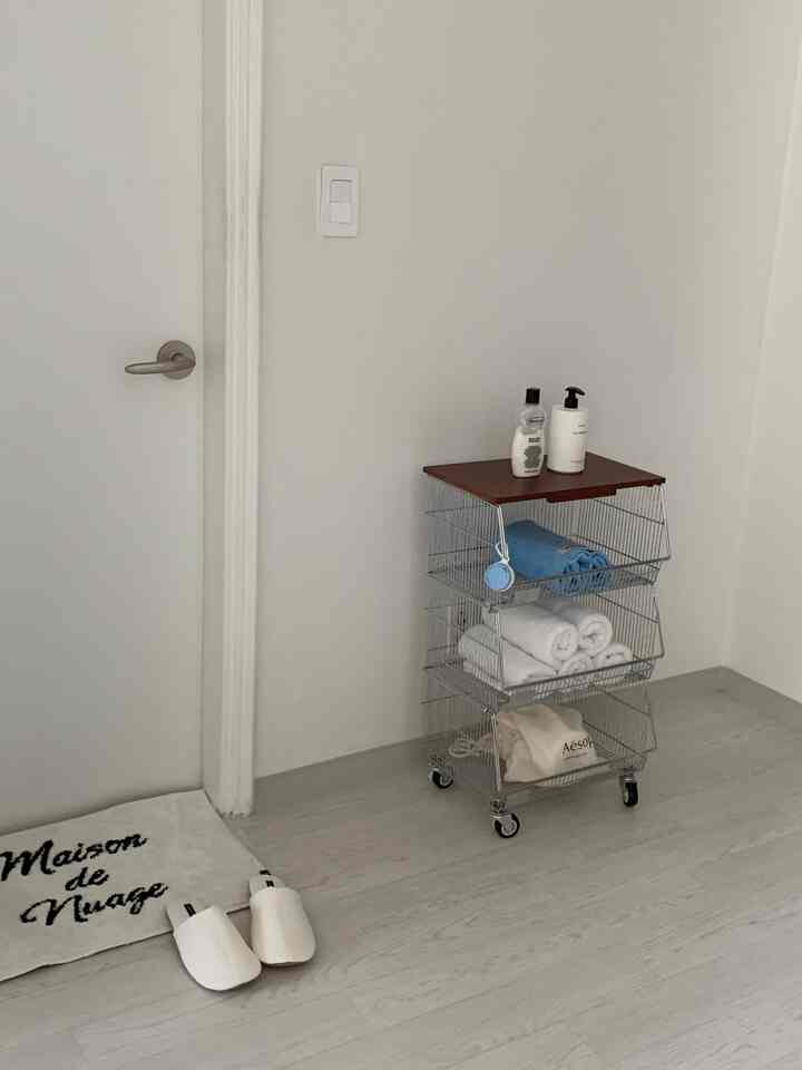 Bright white-toned bathroom area featuring a wheeled 3-tier metal trolley neatly holding towels and body lotion bottles
