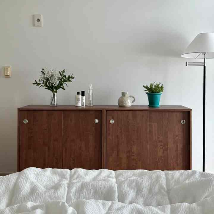 白と茶色のトーンの寝室、引き戸収納キャビネットとフロアランプ、植物鉢や花瓶が配置されたナチュラルなインテリア