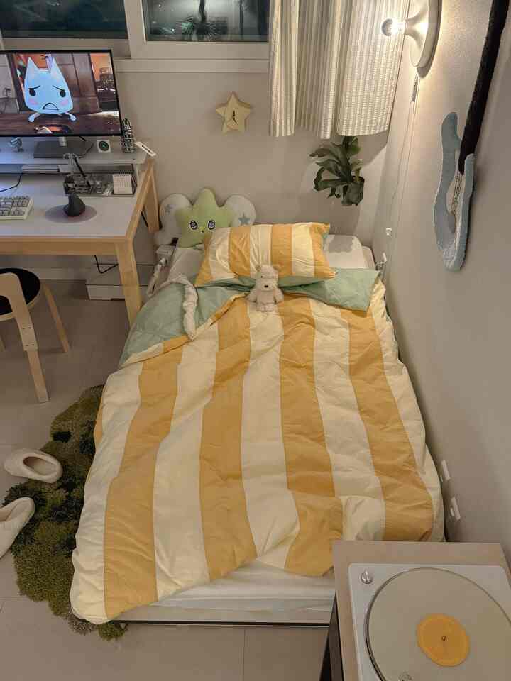 白い壁と黄色のストライプの寝具がある一人用寝室、デスクとコンピュータモニターが配置された落ち着いた空間