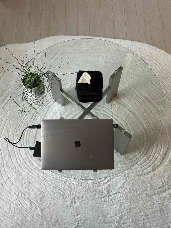 明るいホワイトトーンのカーペットの上に透明な円形ガラステーブルとノートパソコン、植物が配置されたモダンでミニマルなホームオフィス空間