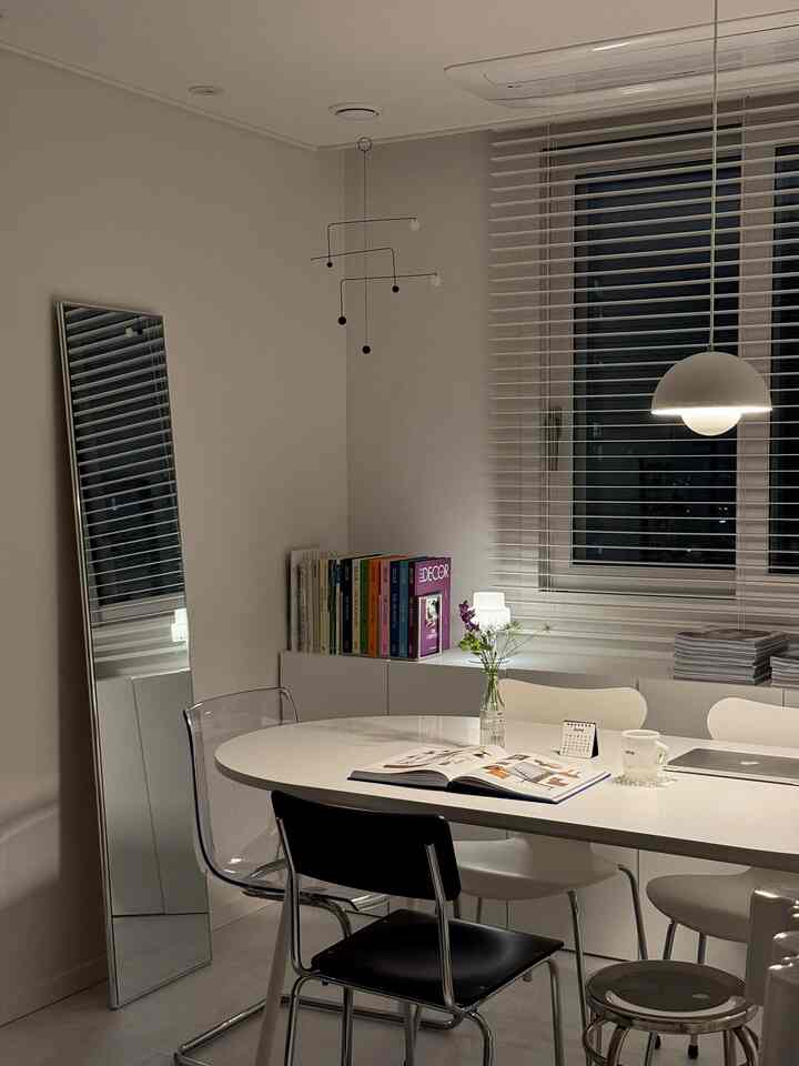A white-toned dining room featuring a central elongated oval table, various chairs, a pendant light, and a mobile, creating a minimalistic space.