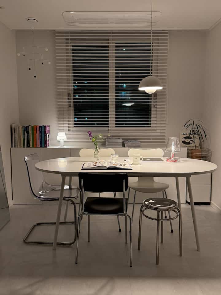Calm white-toned dining room and study featuring oval white dining table, mixed chairs, shelving and blinds with a modern style