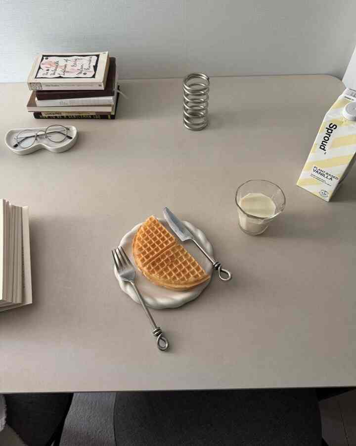 Minimal home cafe scene on beige-toned table featuring waffles, metal cutlery, and glass of milk